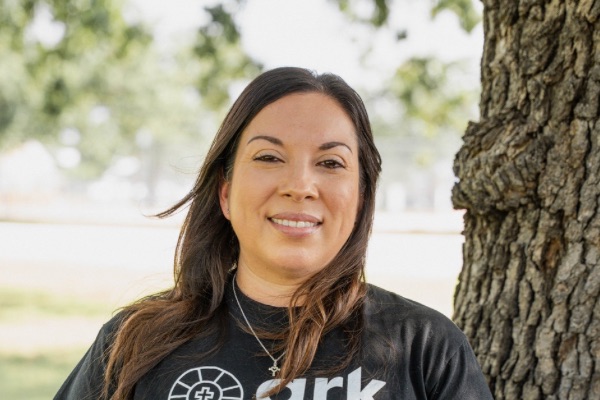 Maggie standing outside of Ark of Highland Lakes offices with black ark logo t-shirt and a tree