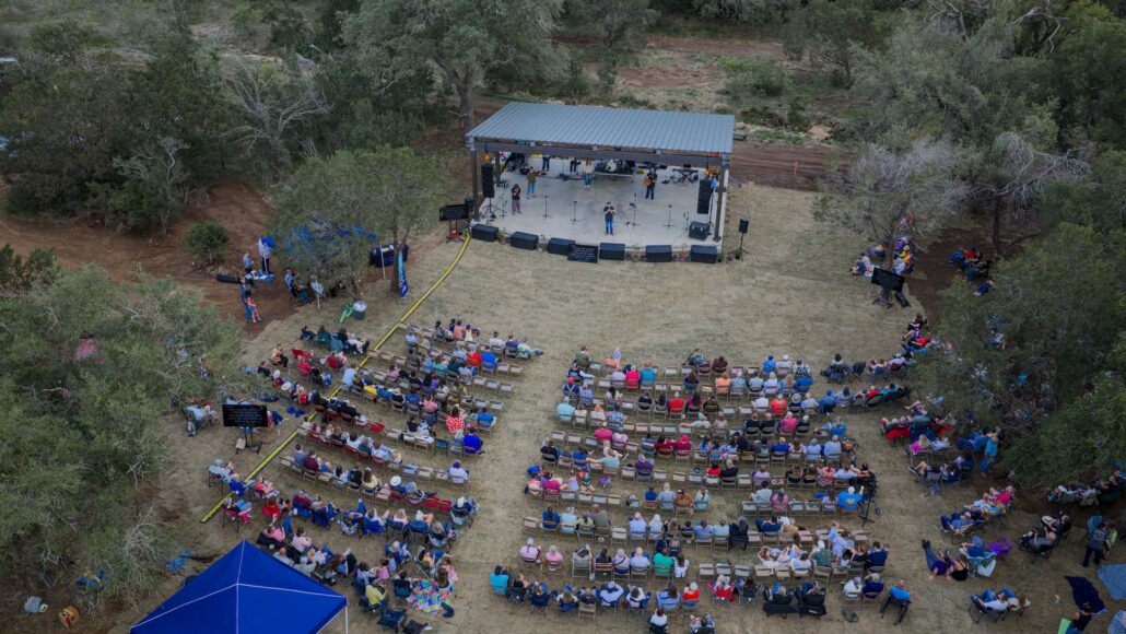 Aerial view of an outdoor music performance on a covered stage in a forest clearing, with rows of audience seating on the grass.