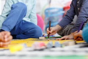Children drawing with pencils on a yellow art table, surrounded by colorful supplies in a bright room, blurred background.