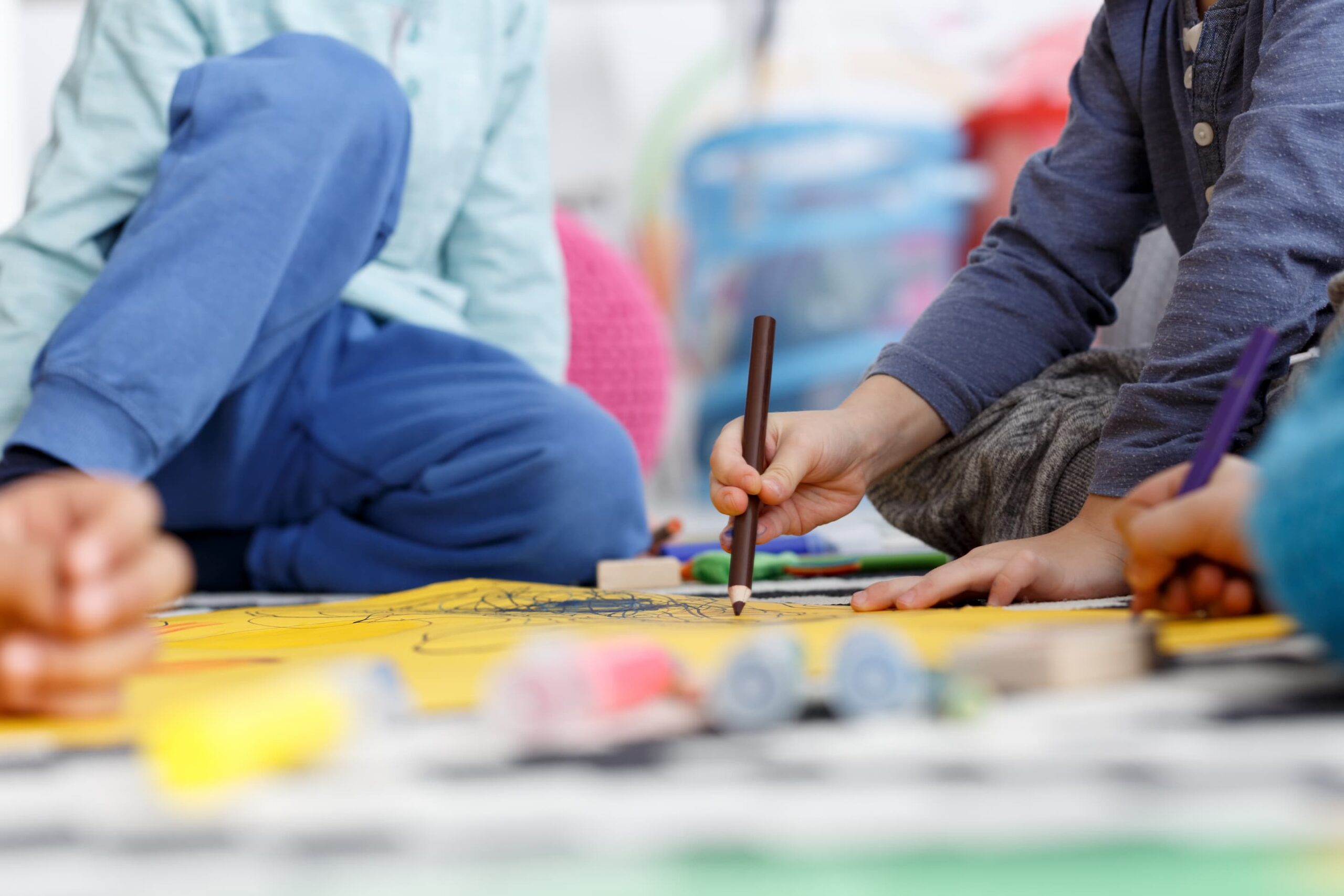 Children drawing with pencils on a yellow art table, surrounded by colorful supplies in a bright room, blurred background.