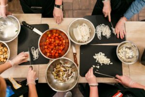Overhead view of people wearing gloves, chopping and preparing ingredients on cutting boards with bowls of tomatoes, noodles, and garlic nearby (cooking class scene).