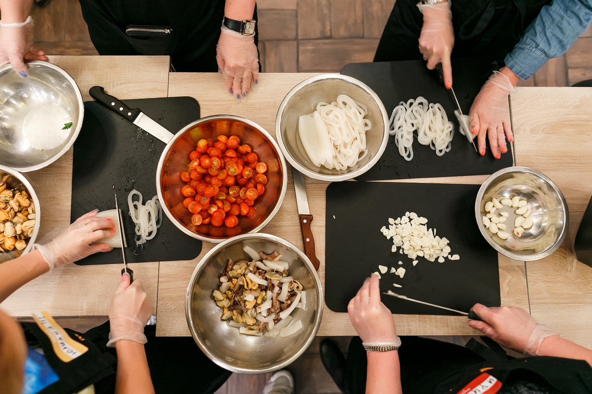 Overhead view of people wearing gloves, chopping and preparing ingredients on cutting boards with bowls of tomatoes, noodles, and garlic nearby (cooking class scene).