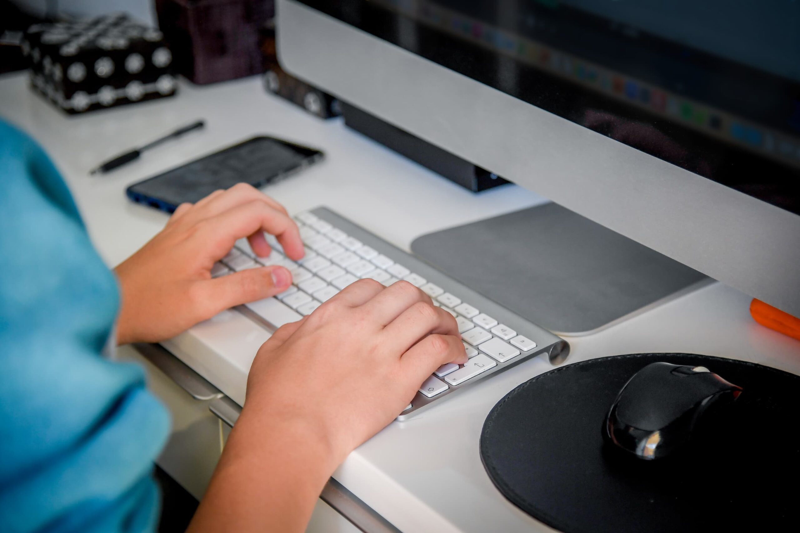 Person typing on a white keyboard at a desk with a monitor and smartphone nearby.