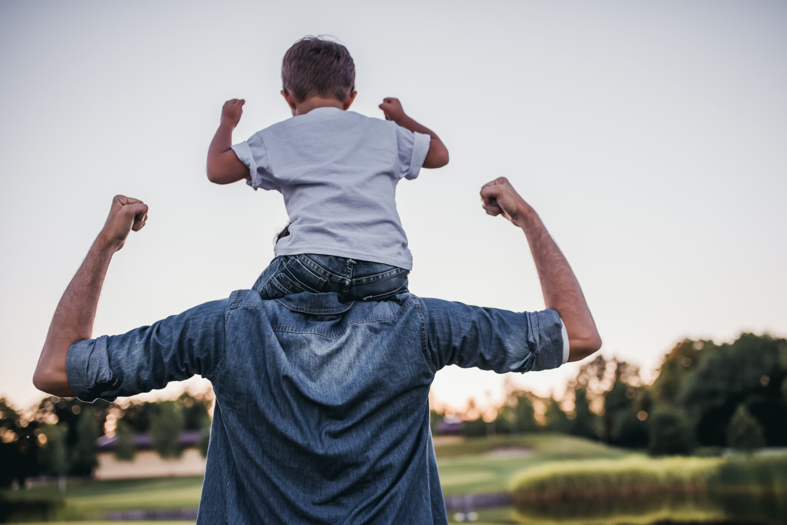 Child sits on an adult's shoulders with arms raised in triumph against a sunset/park backdrop, denim shirt visible at center.