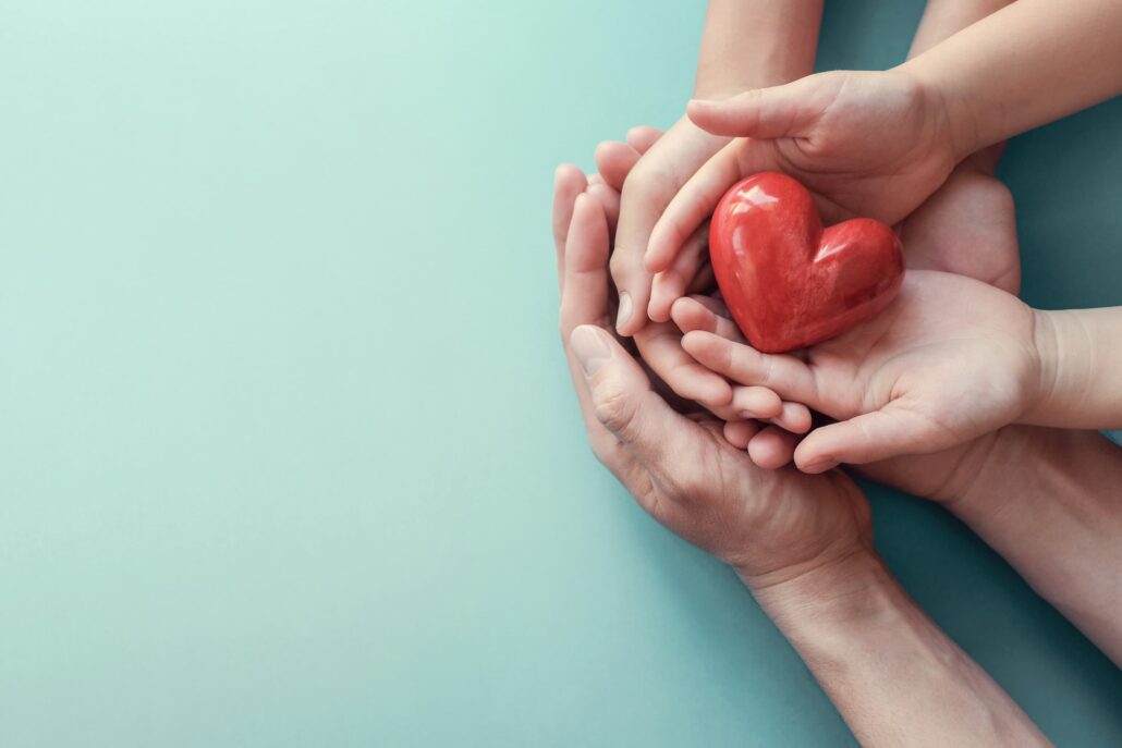 Hands cupping a glossy red heart against a light teal background.