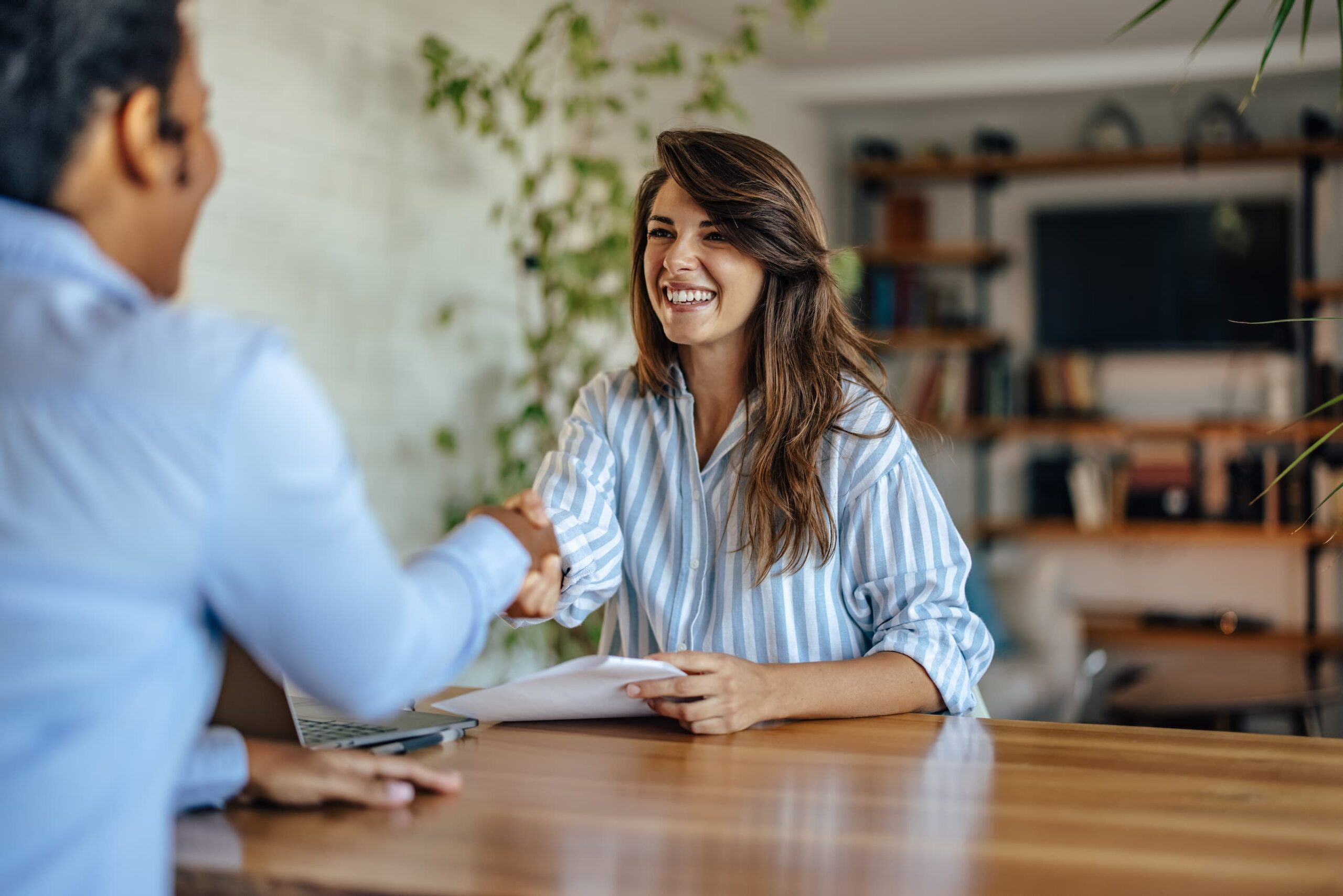 Smiling woman shakes hands with a colleague across a wooden table in a bright office.