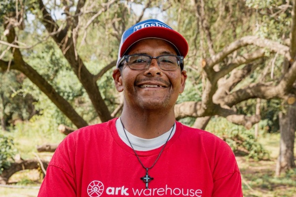 Smiling man wearing a red ark Warehouse t-shirt, glasses, and a necklace with a cross, outdoors with trees in the background.
