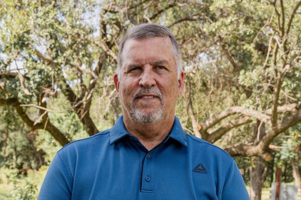 Portrait of a middle-aged man with short gray hair and a beard standing outdoors, wearing a blue polo shirt.