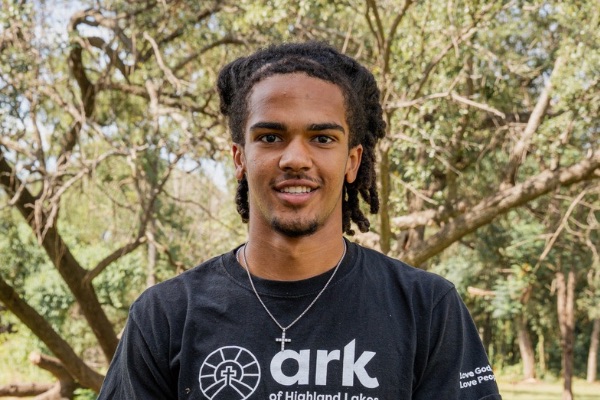 Portrait of a young man with braided hair smiling outdoors, wearing a black T-shirt with a circular logo and 'ark' text.