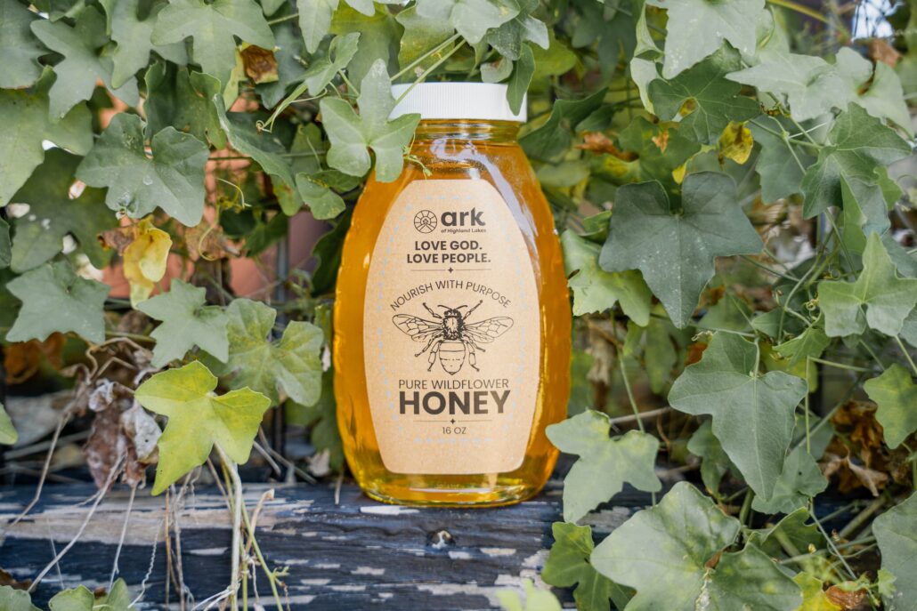 Bottle of honey labeled Ark, nestled among green ivy leaves on a weathered wooden surface.