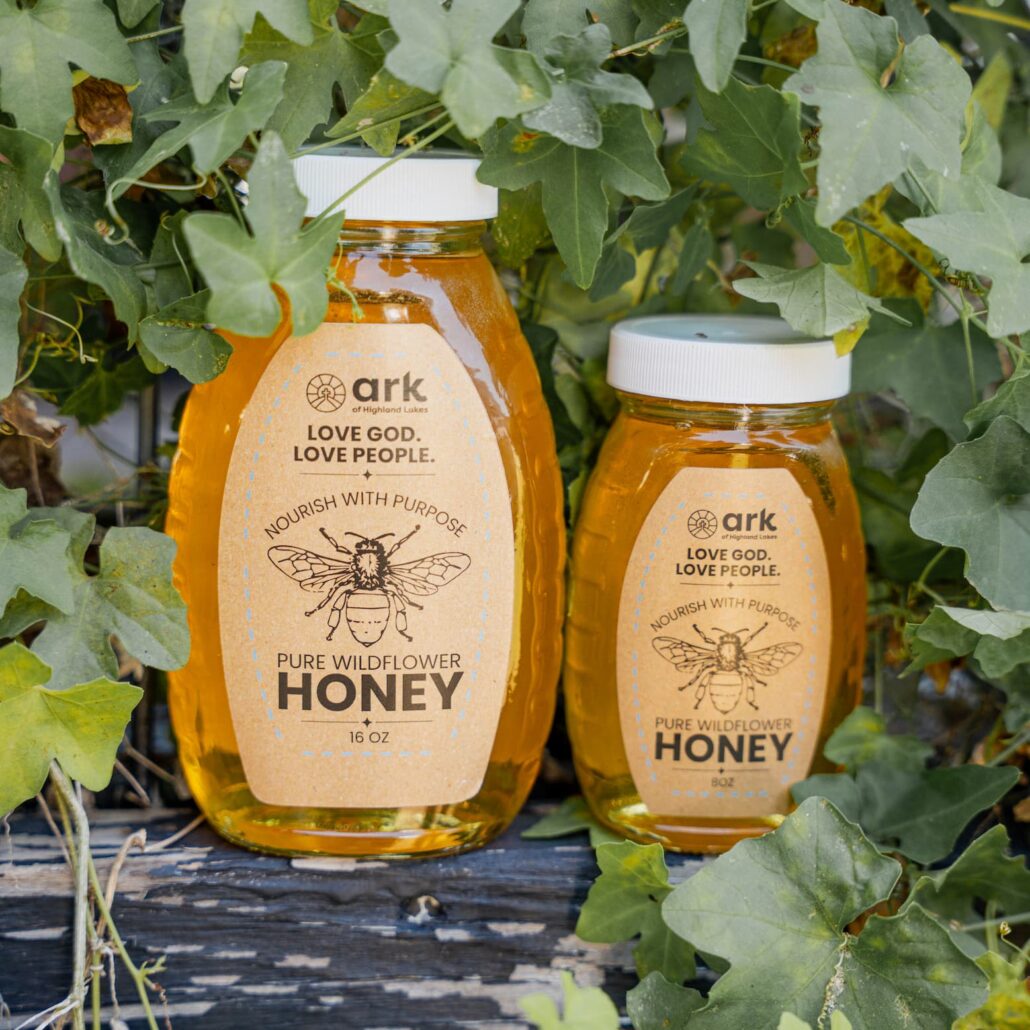 Two honey bottles nestled among ivy leaves, with labels reading 'Love God. Love People. Pure Wildflower Honey.'