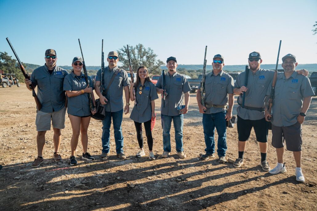 Eight people stand together outdoors, each holding a shotgun, wearing matching gray shirts for a group shooting event on a dirt field.
