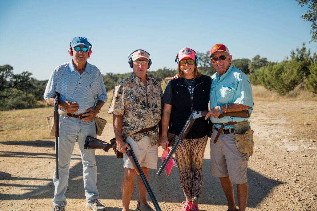 Four adults stand together outdoors at a sunny shooting range, each holding a shotgun and wearing hearing protection and caps/decorative clothing.