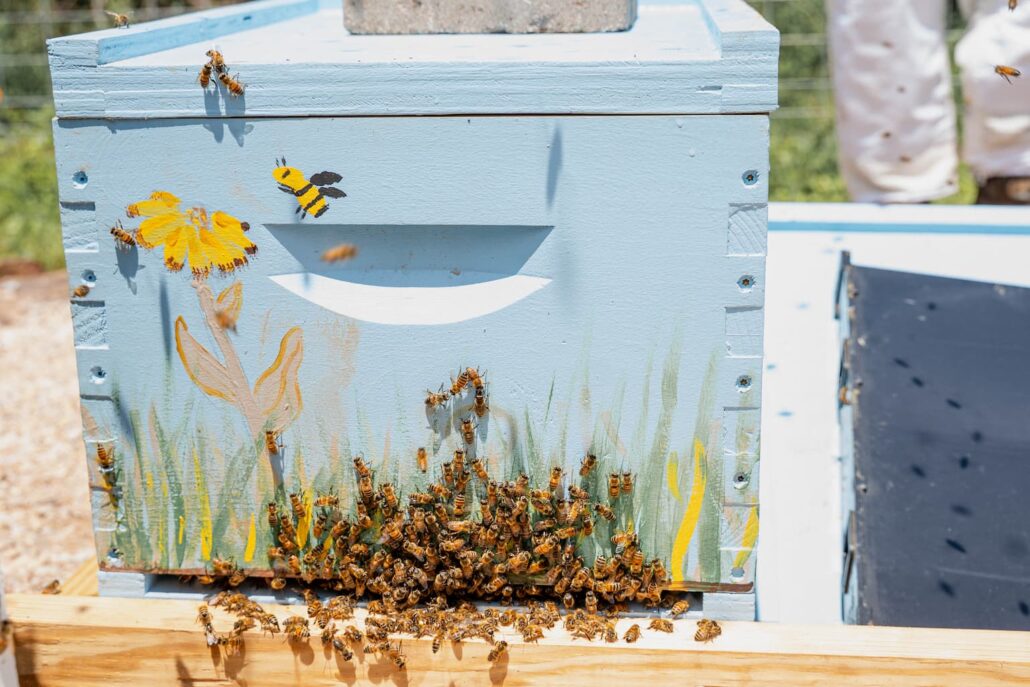 Bees swarm around the entrance of a light blue beehive painted with flowers and grasses.