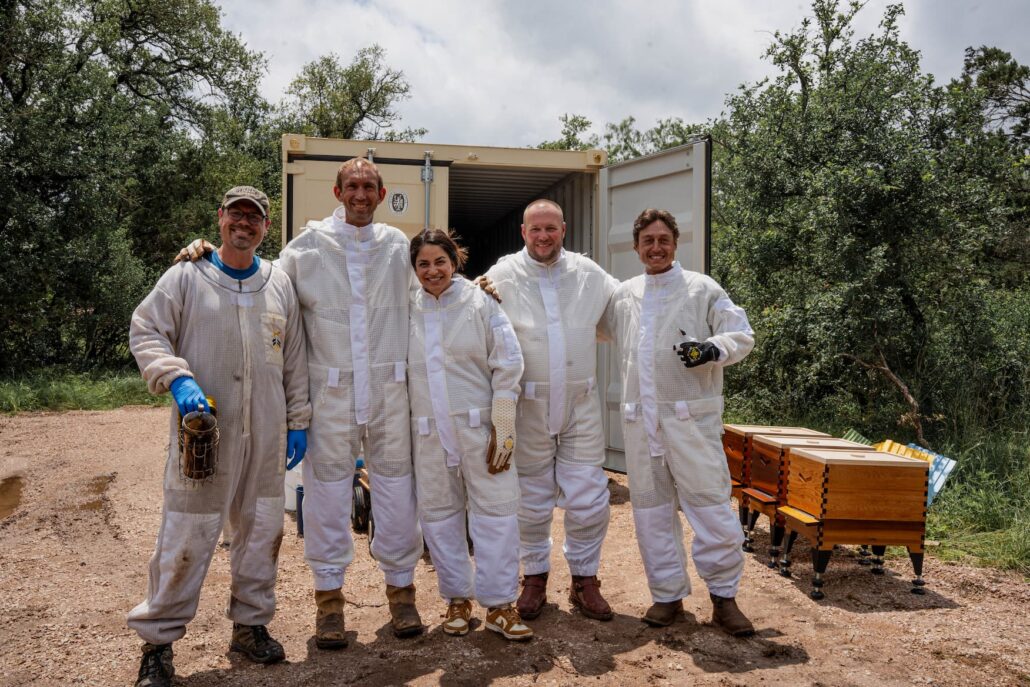 Five beekeepers in white protective suits smile and pose outdoors beside a shipping container and beehives in a rural setting.