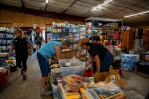 People sorting donated supplies in a cluttered warehouse with shelves full of boxes and products on display beneath a curved ceiling.
