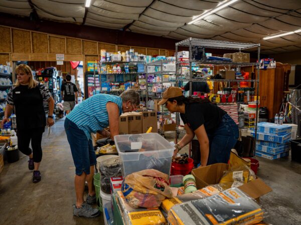 People sorting donated supplies in a cluttered warehouse with shelves full of boxes and products on display beneath a curved ceiling.