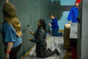 Group of people painting a wall indoors; a kneeling person uses a paint roller while others stand nearby in blue shirts, walls darkened by paint.