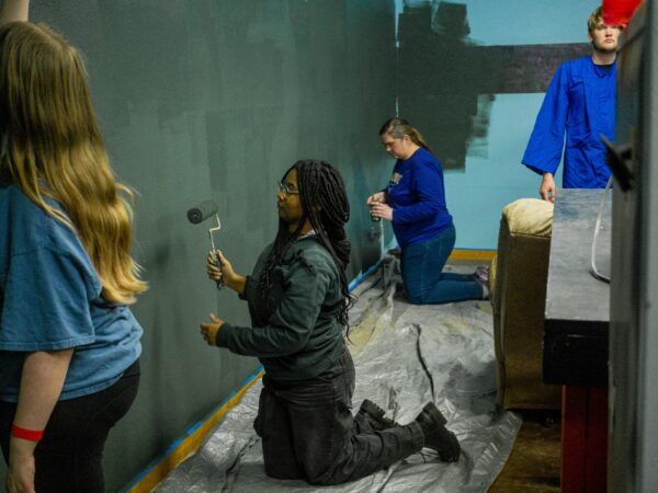 Group of people painting a wall indoors; a kneeling person uses a paint roller while others stand nearby in blue shirts, walls darkened by paint.
