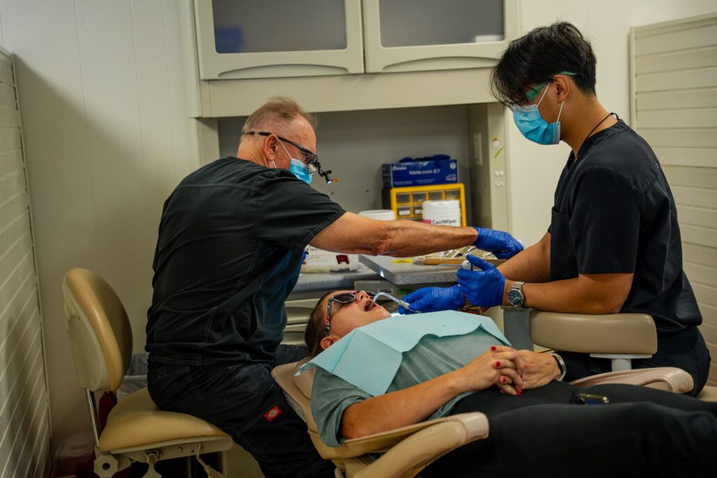 Dentists seated at a dental chair performing a procedure on a patient wearing a bib and mouth opened wide for treatment.