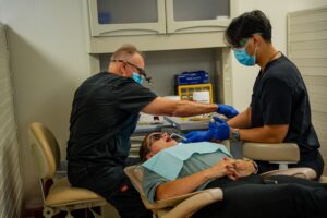 Dentists seated at a dental chair performing a procedure on a patient wearing a bib and mouth opened wide for treatment.