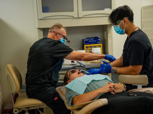 Dentists seated at a dental chair performing a procedure on a patient wearing a bib and mouth opened wide for treatment.