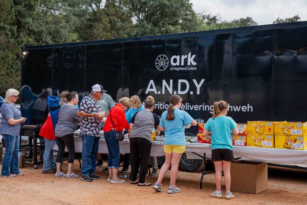 People line up at a food distribution event beside a black truck trailer with 'ARK of Highland Lakes' branding and large 'ANDY' letters, snack boxes in front.