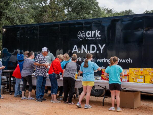 People line up at a food distribution event beside a black truck trailer with 'ARK of Highland Lakes' branding and large 'ANDY' letters, snack boxes in front.