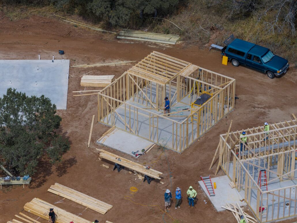 Aerial view of a house under construction with wooden framing, lumber piles, and a blue van on a dirt site at the edge of a wooded area.
