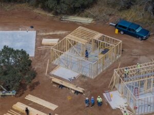Aerial view of a house under construction with wooden framing, lumber piles, and a blue van on a dirt site at the edge of a wooded area.