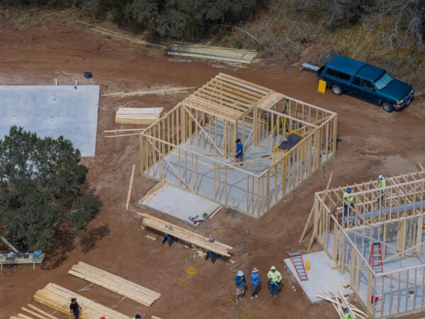 Aerial view of a house under construction with wooden framing, lumber piles, and a blue van on a dirt site at the edge of a wooded area.
