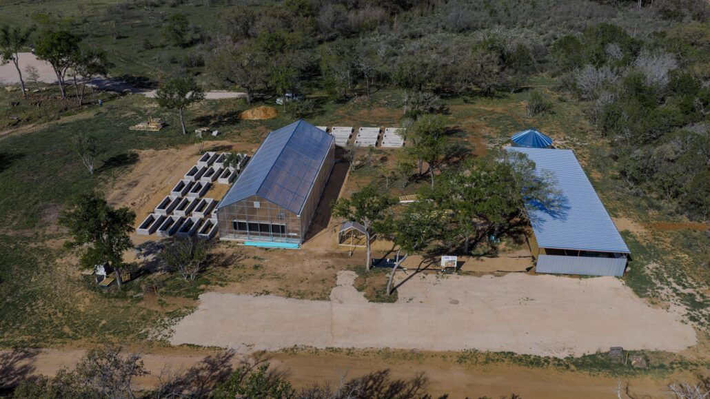 Aerial view of a farm complex with a glass greenhouse, stacked concrete blocks, and a blue-roofed barn among trees appears in a rural clearing.