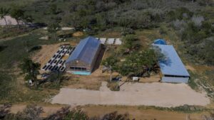 Aerial view of a farm complex with a glass greenhouse, stacked concrete blocks, and a blue-roofed barn among trees appears in a rural clearing.