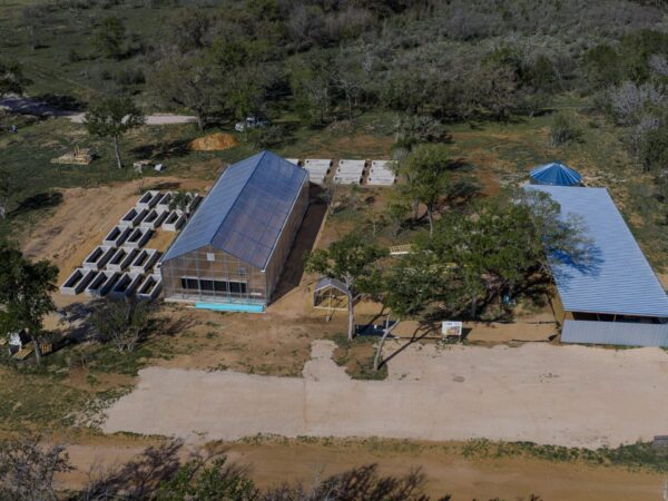 Aerial view of a farm complex with a glass greenhouse, stacked concrete blocks, and a blue-roofed barn among trees appears in a rural clearing.