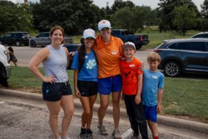 Five people in athletic wear pose together outdoors, smiling in a parking lot with trees and cars behind them.