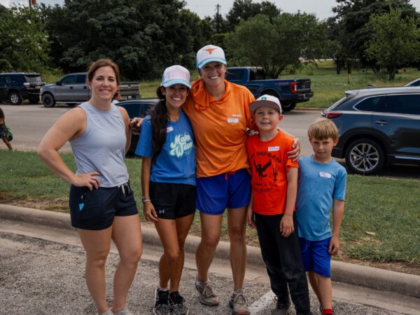 Five people in athletic wear pose together outdoors, smiling in a parking lot with trees and cars behind them.