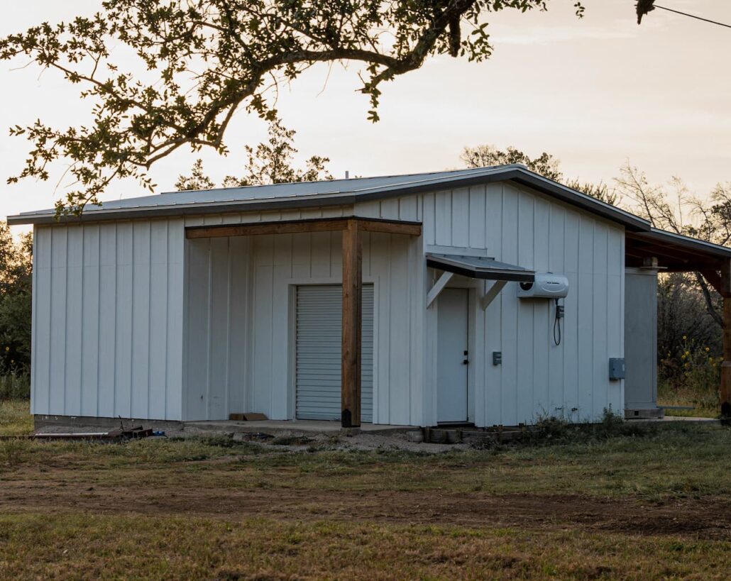 Small white metal utility building in a rural yard at sunset, with a roll-up door and an outdoor AC unit on the side.