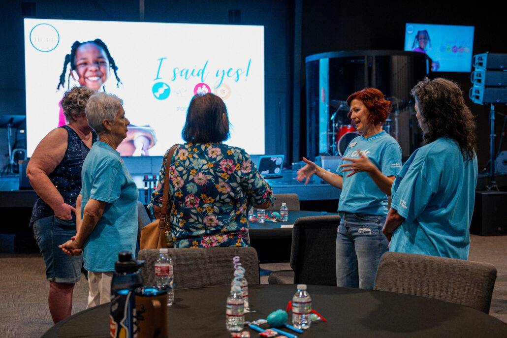 Group of adults in a conference room chat near a round table, with a large screen showing a smiling child and the phrase 'I said yes!' in the background.