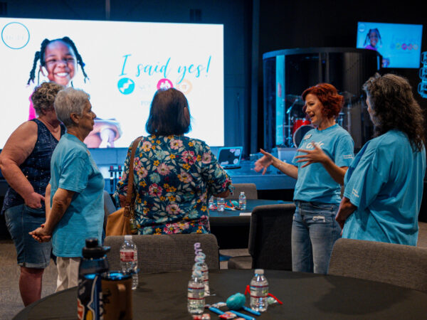 Group of adults in a conference room chat near a round table, with a large screen showing a smiling child and the phrase 'I said yes!' in the background.