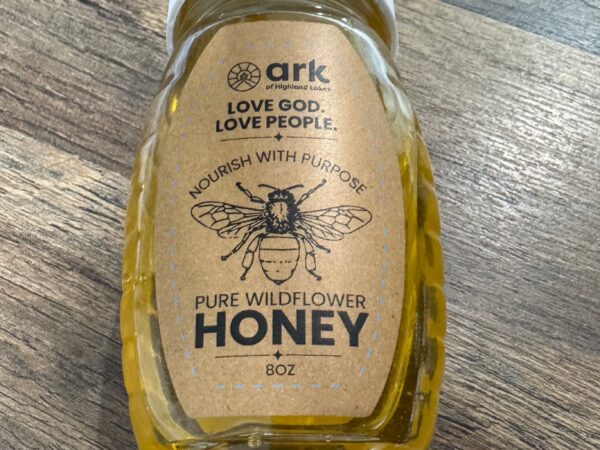Bottle of pure wildflower honey on a wood table; label reads 'Love God, Love People' and 'Pure Wildflower Honey'.