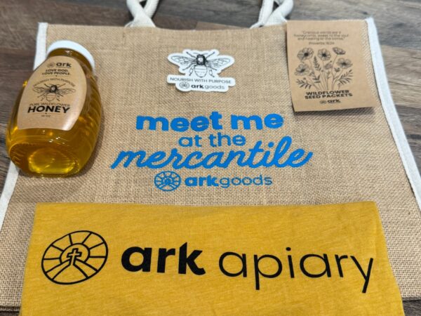 Burlap tote bag with blue 'meet me at the mercantile' logo, a honey bottle on the left, and a wildflower seed packet on the right.