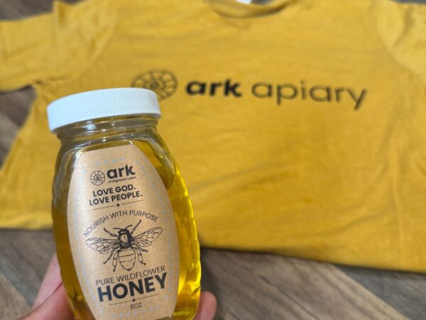 Hand holding a small glass bottle of honey with a white lid; label reads 'Pure Wildflower Honey' and a bee illustration, honey-yellow liquid visible in the bottle.