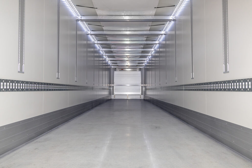 Empty metal trailer interior viewed from the back, with bright LED strip lights along the ceiling and vertical rails on both sides.