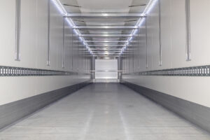 Empty metal trailer interior viewed from the back, with bright LED strip lights along the ceiling and vertical rails on both sides.