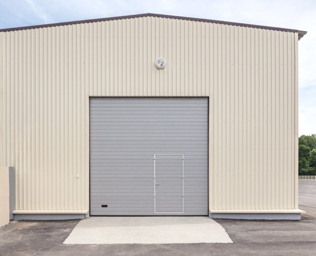 Front view of a beige corrugated metal warehouse with a large gray roll-up door and a round vent above.