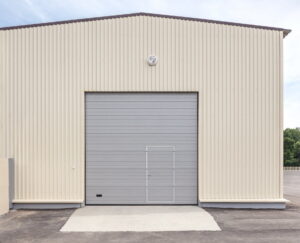 Front view of a beige corrugated metal warehouse with a large gray roll-up door and a round vent above.