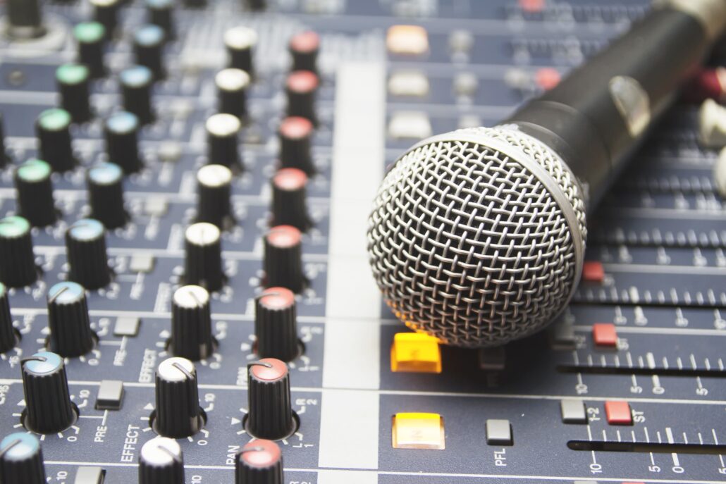Close-up of a silver microphone on a recording mixer with faders and colorful knobs.