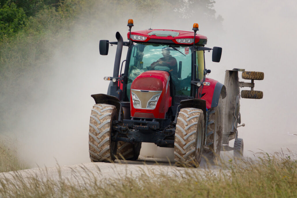 Red agricultural tractor driving toward the camera on a dusty dirt road, kicking up a cloud of dust.