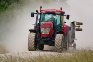 Red agricultural tractor driving toward the camera on a dusty dirt road, kicking up a cloud of dust.
