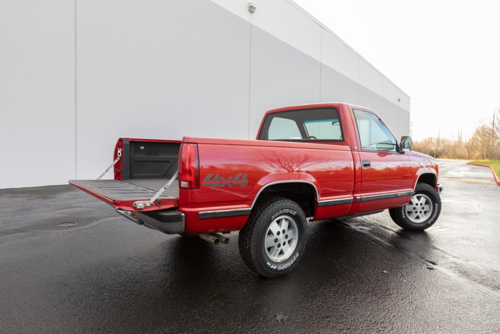 Red pickup truck (4x4) with its tailgate and loading ramp extended, parked beside a gray industrial wall on a wet pavement.